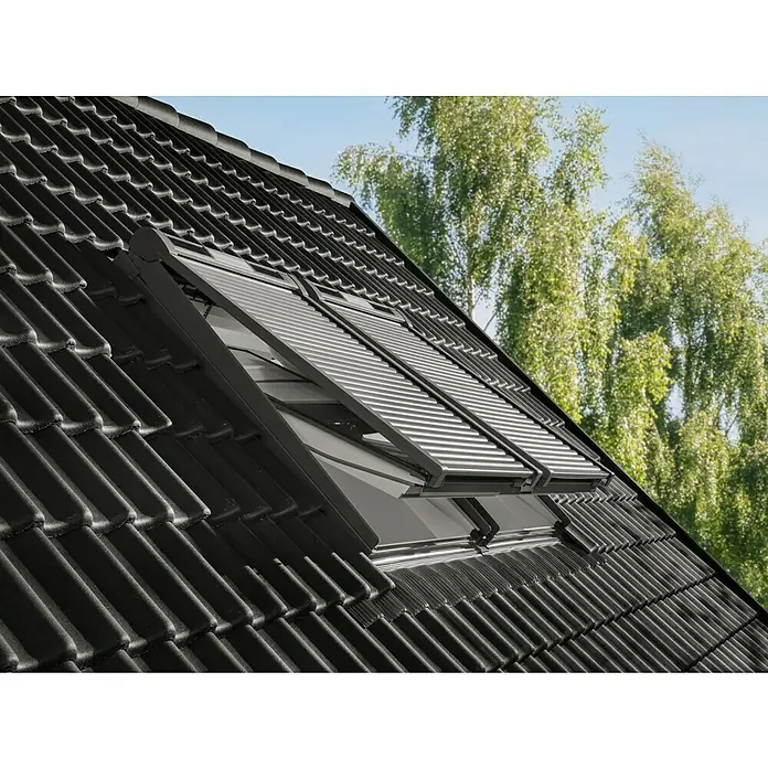 Velux Dachfenster-Rollladen Solar SSL CK06 0000K - Passend für Fenstergröße: CK06, C06, Passend für Fenstertyp: GGL, GGU, GPL, GPU, GHL, GHU, GZL, GLL, GLU, VU und VKU - Zwei dunkle Dachfenster mit teiloffenen Rollläden auf schwarzem Ziegeldach, Bäume und Himmel im Hintergrund.
