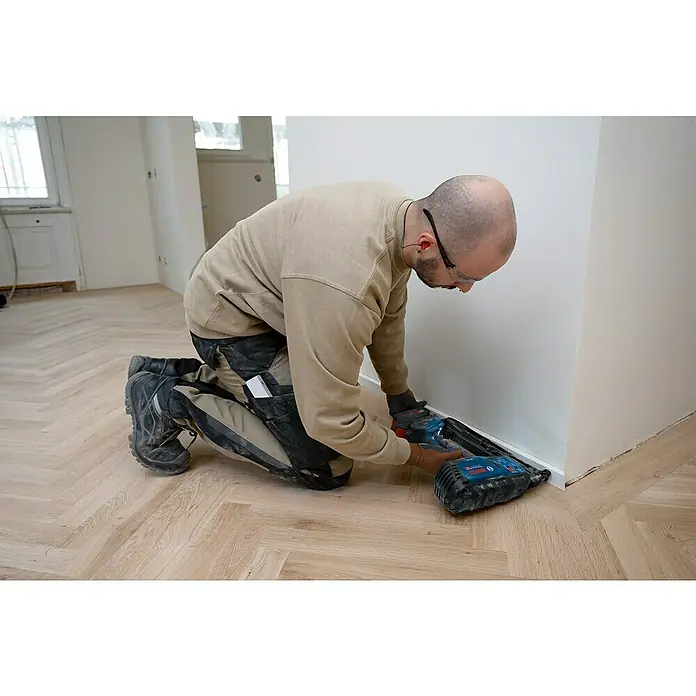 Bosch Professional Akku-Nagler GNH 18V-64-2 M PRO SOLO - 18 V, Ohne Akku, Länge Nagel: 32 mm - 64 mm - Mann verwendet eine blau-schwarze Bosch Nagelpistole zur Montage einer weißen Fußleiste auf einem hellen Holzboden.