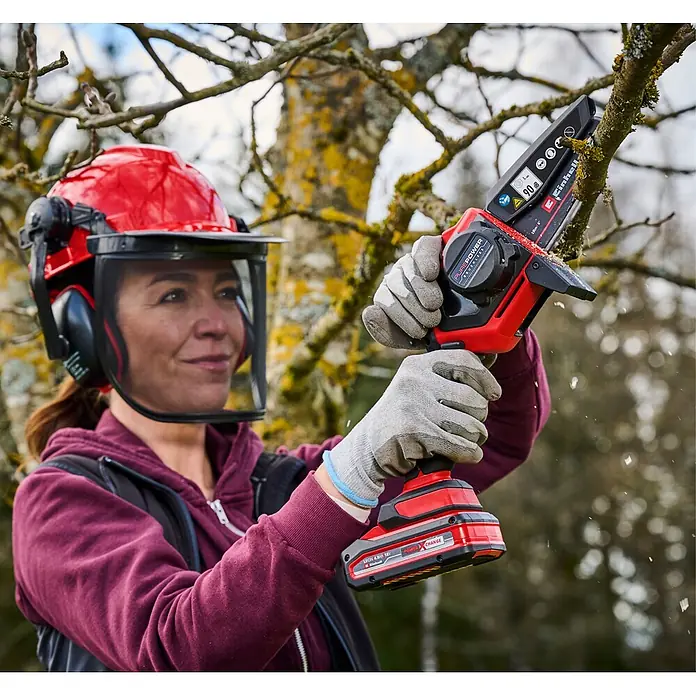 Einhell Power X-Change 18V Einhand-Akku-Kettensäge GE-PS 18/15 Li Holster Kit - 18 V, Li-Ionen, Ohne Akku, Schwertlänge: 15,8 cm - Frau mit Helm und Handschuhen benutzt eine rot-schwarze Akku-Minikettensäge zum Baumschnitt.