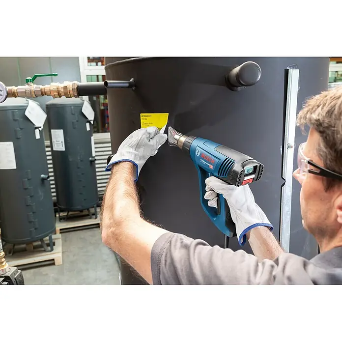 Bosch Professional Heißluftgebläse GHG 23-66 PR - Temperaturbereich: 50 °C - 650 °C - Eine Person mit Handschuhen und Schutzbrille nutzt eine blau-schwarze Bosch Professional Heißluftpistole, um ein gelbes Etikett auf einem großen schwarzen Tank zu erhitzen.