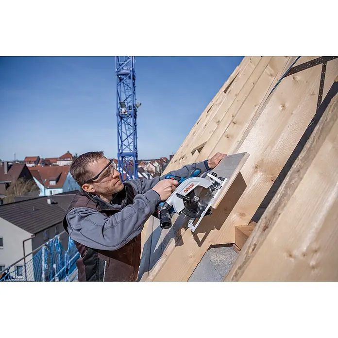 Bosch Professional Akku-Handkreissäge - 18 V, 2 Akkus, Durchmesser Sägeblatt: 165 mm, 5.000 U/min - Mann benutzt eine blau-silberne Bosch Handkreissäge zum Schneiden eines Holzbretts auf einem Dachstuhl.