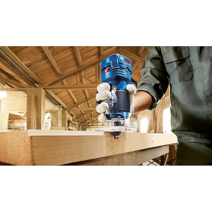 Bosch Professional Elektro-Oberfräse GLF 55-6 - 550 W, 33.000 U/min - Eine Person fräst mit einer blauen Bosch Professional Oberfräse und Handschuh ein helles Holzstück in einer Werkstatt.