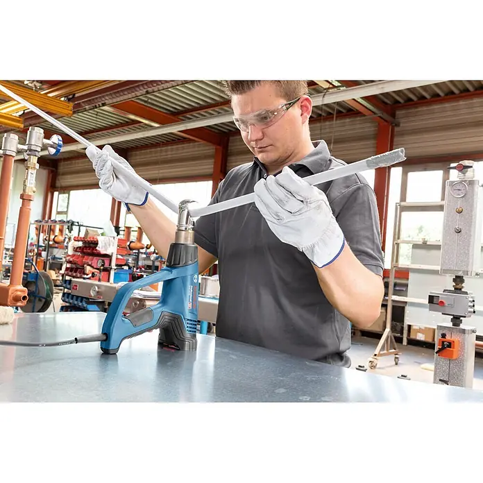 Bosch Professional Heißluftgebläse GHG 23-66 PR - Temperaturbereich: 50 °C - 650 °C - Blau-schwarze Bosch Heißluftpistole steht auf Metalltisch, erwärmt ein helles Rohr, das von einer Person mit Schutzbrille und Handschuhen gebogen wird.