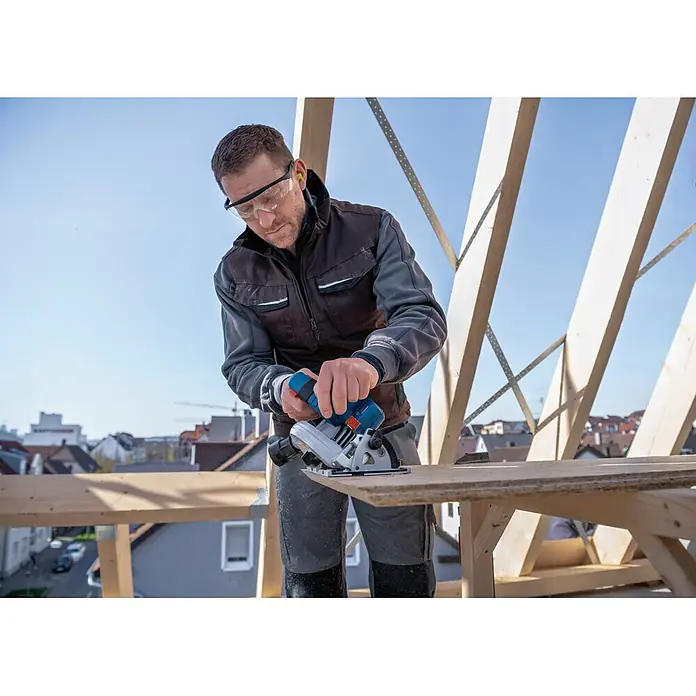 Bosch Professional Akku-Handkreissäge - 18 V, 2 Akkus, Durchmesser Sägeblatt: 165 mm, 5.000 U/min - Mann benutzt eine blaue und silberne Kompaktkreissäge, um Holz auf einer Baustelle zu sägen, mit Sicherheitsausrüstung.