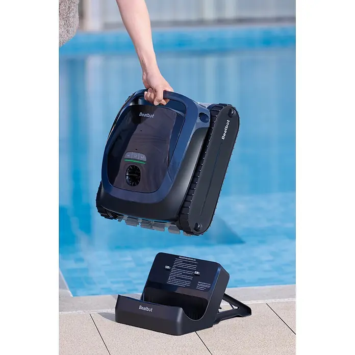 Beatbot Poolroboter AquaSense 2 - Akkubetrieben, Passend für: Pools bis 300 m² Grundfläche, Filterleistung: 20.800 l/h - Eine Hand hält einen dunkelblauen Beatbot Roboter-Poolreiniger mit Kettenlaufwerk über seiner schwarzen Ladestation neben einem blauen Pool.