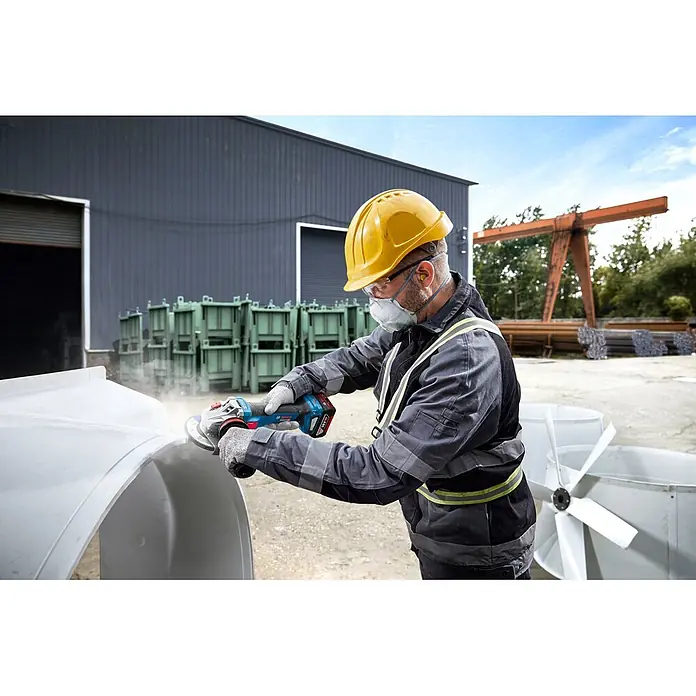 Bosch Professional AMPShare 18V Akku-Winkelschleifer GWS 18V-11 S PRO - Ohne Akku, Durchmesser Scheibe: 125 mm - Eine Person mit Schutzkleidung schleift ein großes weißes Industrieteil mit einem blauen Bosch Akku-Winkelschleifer, wodurch Staub entsteht.