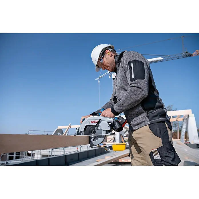 Bosch Professional AMPShare 18V Akku-Handkreissäge GKS 18V-70 L PRO - 18 V, 2 Akkus, Durchmesser Sägeblatt: 190 mm, 5.000 U/min - Mann in Schutzkleidung schneidet mit einer silber-schwarzen Bosch Akku-Kreissäge einen Holzbalken auf einer Baustelle.