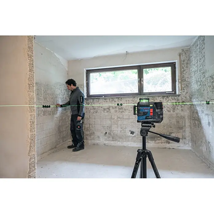 Bosch Professional Linienlaser GLL 12V-100-33 CG PRO - Max. Arbeitsbereich: 30 m - Blau-schwarzes Bosch Professional Laser-Nivelliergerät auf Stativ projiziert eine grüne Linie, die ein Mann zur Wandausrichtung bei einer Renovierung nutzt.