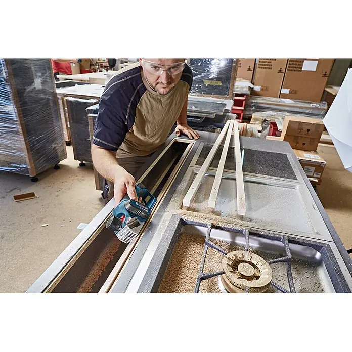 Bosch Professional Akku-Hobel GHO 12V-20 PRO - 12 V, 2 Akkus, Hobeltiefe: 0 mm - 2 mm, Leerlaufdrehzahl: 14.500 U/min - Mann hobelt mit blau-schwarzem Bosch Akku-Hobel eine Holzplatte, Späne fallen.