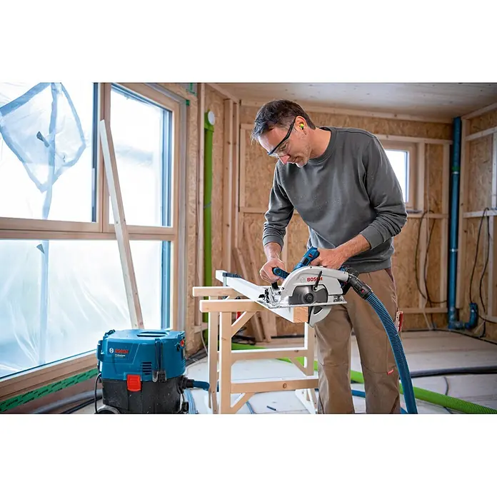 Bosch Professional AMPShare 18V Akku-Handkreissäge GKS 18V-70 L PRO - 18 V, 2 Akkus, Durchmesser Sägeblatt: 190 mm, 5.000 U/min - Mann schneidet Holz mit einer grau-blauen Bosch Handkreissäge, verbunden mit einem blau-schwarzen Bosch Sauger.