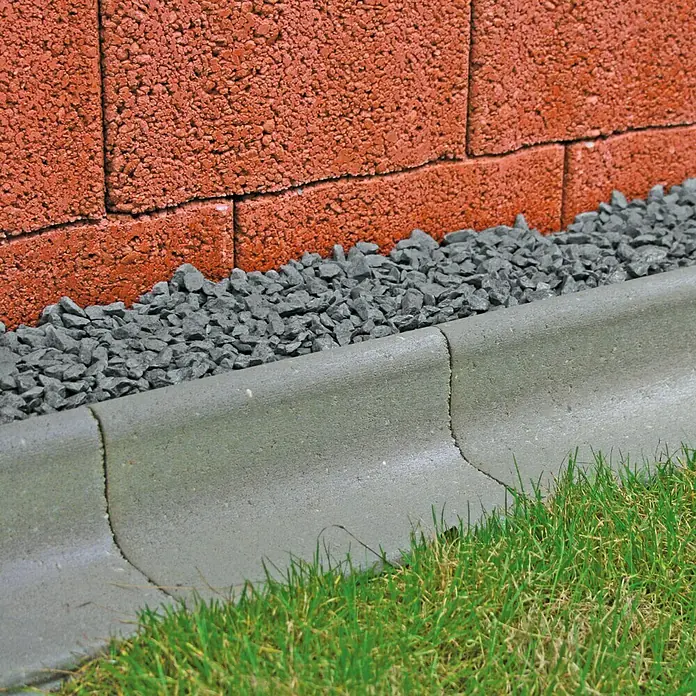 Christoph Mähstein Mährandstein - 25 x 18,5 x 12 cm, Grau, Beton - Nahaufnahme einer grauen Betonbordsteinkante, die einen Gartenweg von einem Rasen trennt, mit Ziegelmauer im Hintergrund.