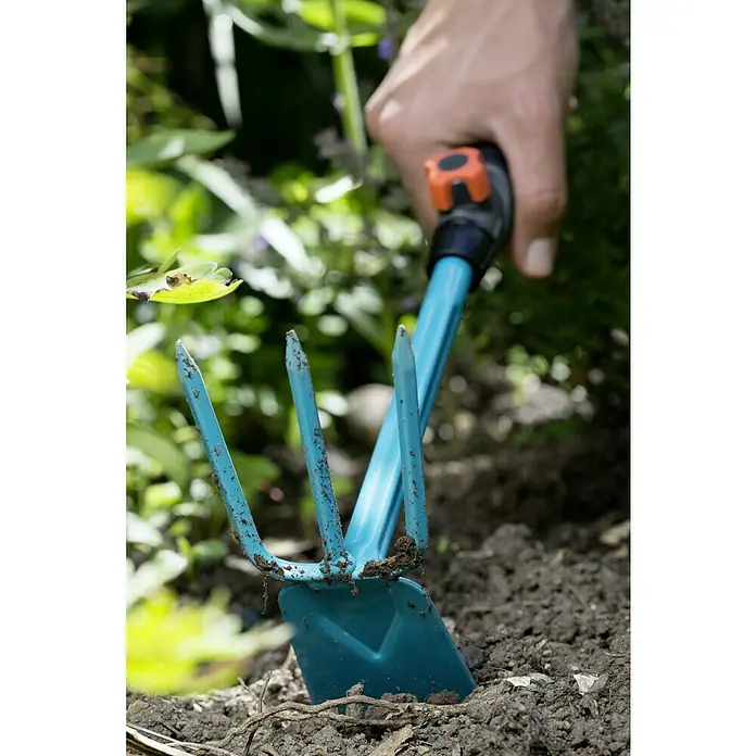 Gardena Combisystem Kleinhacke - Arbeitsbreite: 6,5 cm, Blattform: Gerade, Anzahl Zinken: 3 - Nahaufnahme eines Gartenwerkzeugs, einer blauen Handhacke mit Zinken, die in der Erde steckt, gehalten von einer Hand.