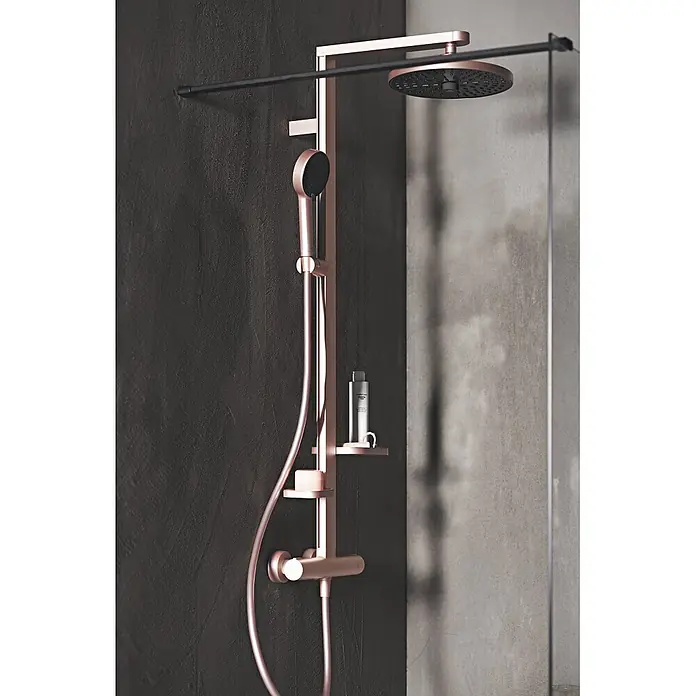 Ideal Standard Alu+ Duschsystem Ceratherm - Anzahl Strahlarten: 2 Stk., Rosé - Kupferfarbenes Duschsystem mit großem Regenduschkopf, Handbrause und zwei Ablagen, an einer dunklen, strukturierten Wand montiert.