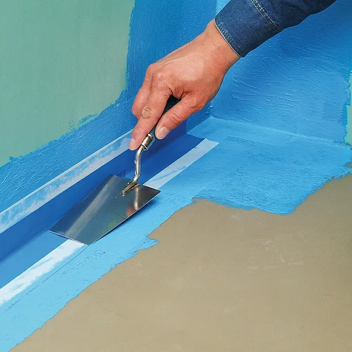 Knauf Flächendichtband - 50 m - Der Handwerker glättet die blaue Dichtmasse mit einer Glättkelle auf dem Boden, um eine wasserdichte Abdichtung zu schaffen.