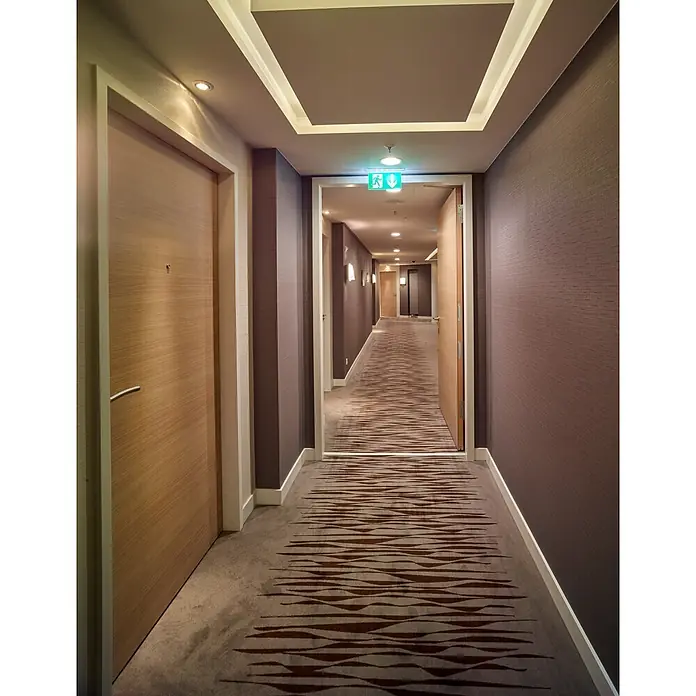 Hörmann Stahlzarge - 875 x 2.000 mm, Wandstärke: 100 mm - Ein moderner Hotelkorridor mit strukturierten braunen Wänden, hellen Holztüren, Einbauleuchten und einem gemusterten Teppichboden, der zu einem Ausgangsschild führt.