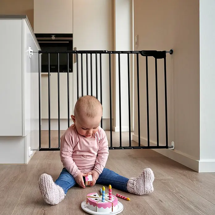 Dolle Sicherheitsgitter Lars - Höhe: 68 cm, Verstellbar: 74,4 cm - 113 cm, Anthrazit - Schwarzes Baby-Schutzgitter in einem Durchgang installiert, das den Zugang zu einer Küche blockiert, wo ein Baby sicher spielt.
