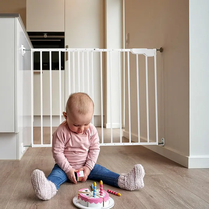 Dolle Sicherheitsgitter Lars - Höhe: 68 cm, Verstellbar: 74,4 cm - 113 cm, Weiß - Weißes Baby-Schutzgitter in einer Türöffnung, das ein auf dem Boden spielendes Baby vom Küchenbereich trennt.