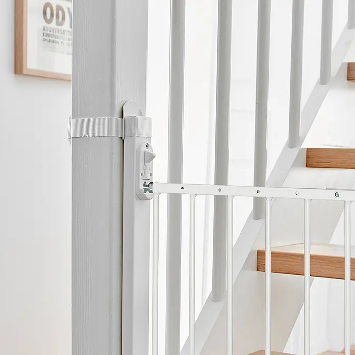 Dolle Sicherheitsgitter Adapter - Weiß - Weißes Metall-Schutzgitter, mit Gurt und Riegel an einem weißen Treppenpfosten befestigt, blockiert eine Treppe.