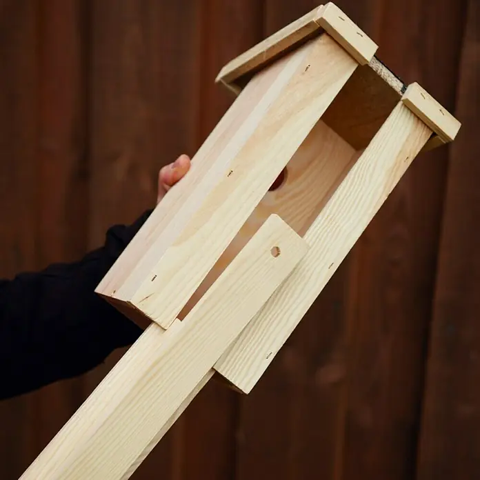 Nistkasten - 17 x 18 x 40 cm, Hellbraun - Nahaufnahme eines handgefertigten Vogelhauses aus Holz mit einem langen Pfosten, das vor einem braunen Holzzaun gehalten wird.