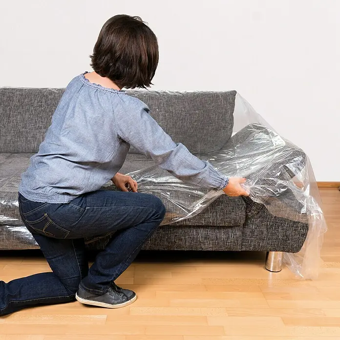 Couchhülle - 300 x 130 cm, Transparent, 2 Stk. - Frau legt eine Schutzfolie über ein Sofa. Ideal, um Möbel während Renovierungsarbeiten oder beim Umzug sauber und geschützt zu halten.