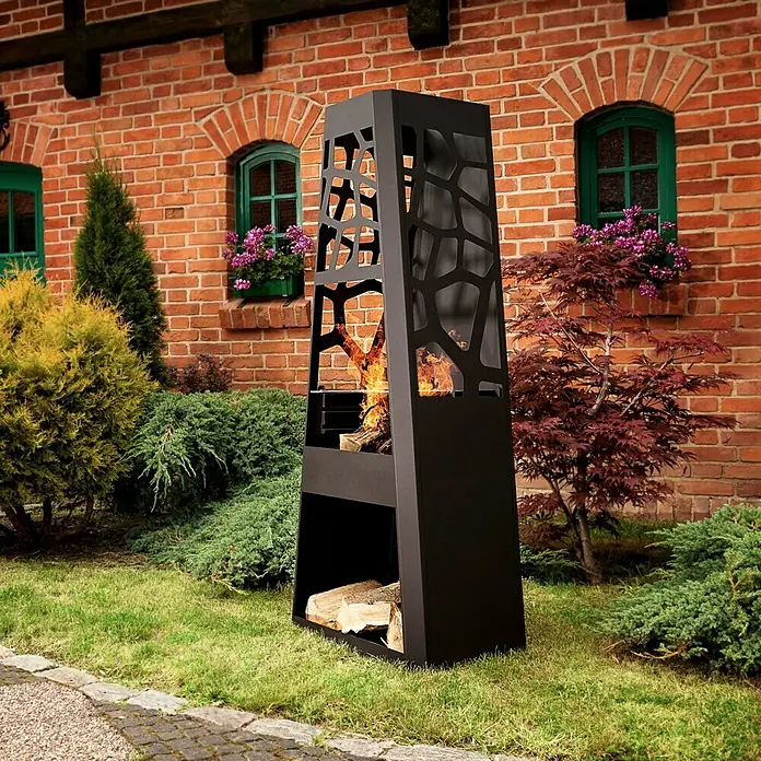 Dobar Gartenkamin - 57,5 x 38 x 148 cm, Stahl, Pulverbeschichtet - Ein moderner, schwarzer Kaminofen mit Holzscheiten steht vor einem roten Backsteingebäude, umgeben von üppigem Grün.