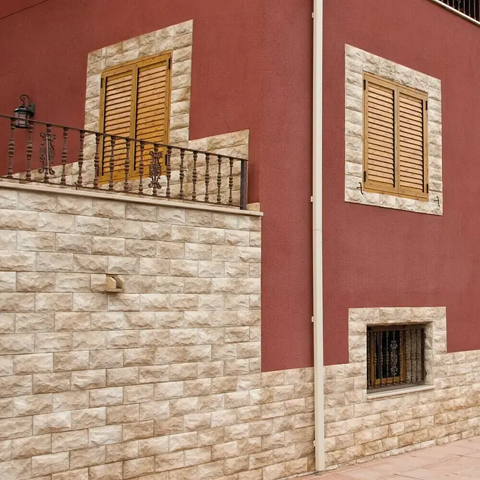 Revestimiento de piedra Teide - 14 x 28 cm, Pardo - Fachada de un edificio con paredes rojas, revestimiento de piedra y contraventanas de madera. La mampostería es decorativa y estructurada.
