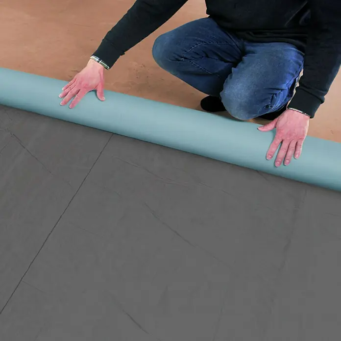 Karibu Premium Dachbahn - Silbergrau, Selbstklebend - Ein Mann kniet, während er eine Rolle hellblauen Vinylboden auf einem grauen Untergrund ausrollt, um einen neuen Bodenbelag zu verlegen.