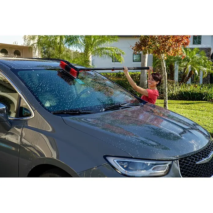 Starlyf Reinigungsbürste Hydrocleaner - Länge Stiel: 4,6 m, Kunststoff, Aluminium - Lächelnde Frau wäscht ein dunkelgraues Auto mit einer rot-schwarzen Waschbürste.