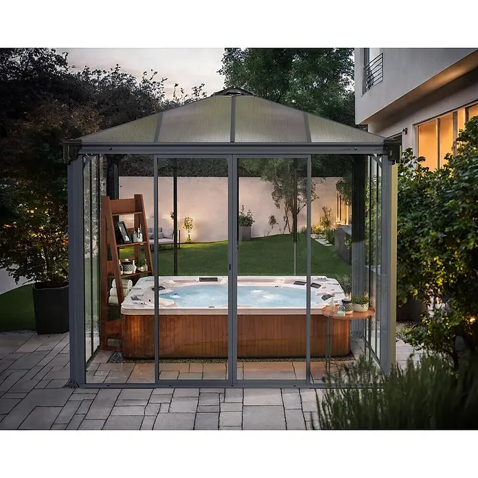 Palram – Canopia Pérgola de jardín cerrado Ledro - An x Pr: 295 x 295 cm, Color del armazón: Antracita, Aluminio - Cerramiento exterior con marco gris y techo translúcido que alberga un jacuzzi burbujeante en un patio ajardinado.