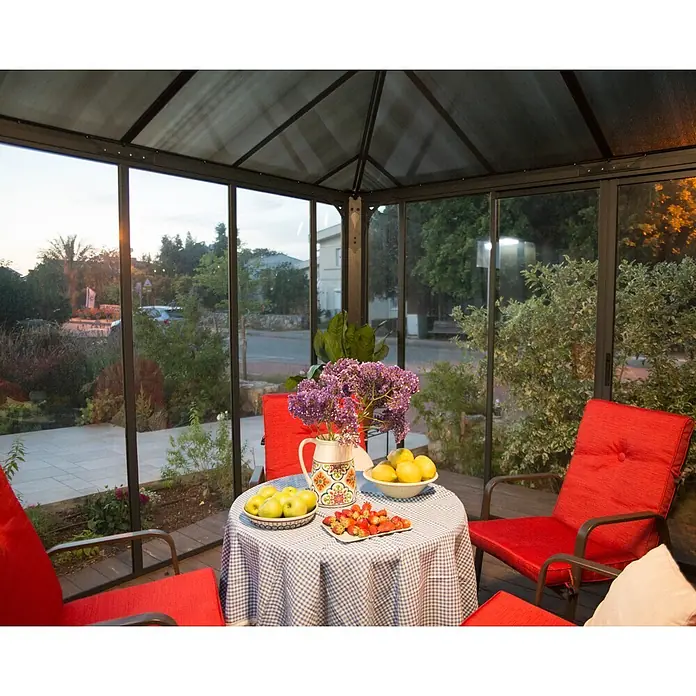 Palram – Canopia Pérgola de jardín cerrado Ledro - An x Pr: 295 x 295 cm, Color del armazón: Antracita, Aluminio - Un cenador de techo transparente con estructura oscura y paredes de cristal, amueblado con sillas rojas y una mesa con fruta.