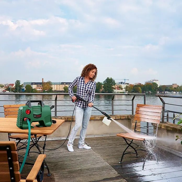 Bosch Hochdruckreiniger EasyAquatak 120 - 1.500 W - Eine Frau benutzt einen grünen Bosch Hochdruckreiniger, um einen Holzstuhl auf einer Terrasse mit Blick auf einen See zu reinigen.