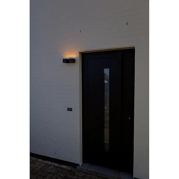Lutec Aplique para exterior LED Fari - 10 W, L x An x Al: 92 x 65 x 193 mm, Gris oscuro, Aluminio, IP54 - Puerta principal moderna negra con inserto de vidrio vertical, flanqueada por una lámpara de pared y un timbre en una pared de ladrillo claro.