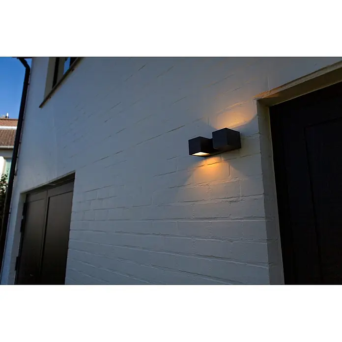 Lutec Aplique para exterior LED Fari - 10 W, L x An x Al: 92 x 65 x 193 mm, Gris oscuro, Aluminio, IP54 - Aplique de pared exterior cúbico gris oscuro en una pared de ladrillo blanca, emitiendo luz cálida arriba y abajo.