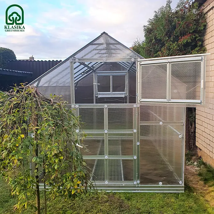 Klasika Greenhouses Gewächshaus Bernard Klasika - Außenmaß inkl. Dachüberstand (B x T): 235 x 600 cm, Material Seitenwand: Kunststoff, 6 mm, Silbergrau - Gewächshaus mit klaren Wellplatten und silbernem Rahmen, mit geöffneter Tür und Lüftung, in einem Garten.