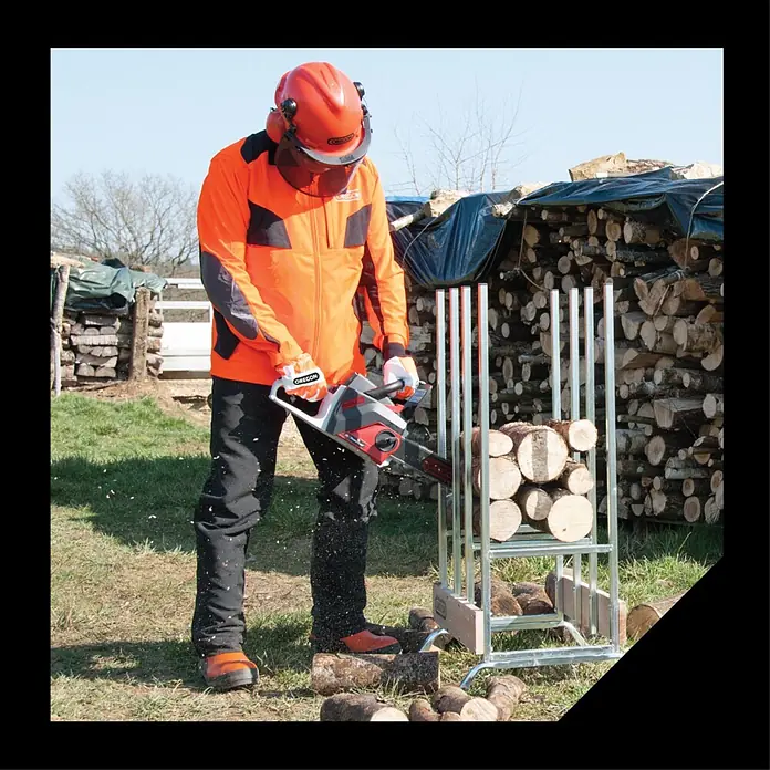 Oregon Pantalones resistentes al corte Yukon - S, Negro/Naranja, Unisex - Persona con equipo de seguridad corta troncos con una motosierra eléctrica gris y roja en un soporte metálico al aire libre.