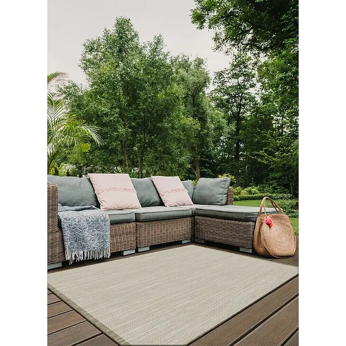 Alfombra exterior reversible In/Out Niebla - Beige, 230 x 160 cm, Polipropileno - Una alfombra de exterior tejida color beige grisáceo sobre una tarima de madera bajo un sofá de ratán con cojines.