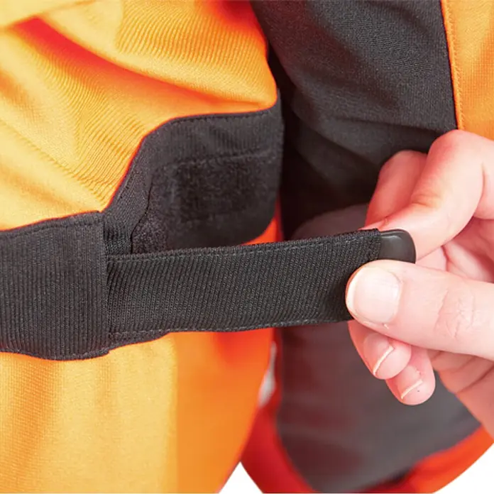 Oregon Chaqueta de trabajo Yukon - XXXL, Naranja/Gris - Una mano ajusta una correa de tela negra con una lengüeta de plástico en una prenda naranja y negra, indicando ajuste.