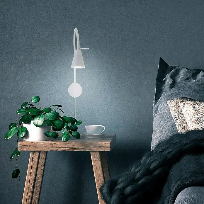 Briloner Foco de pared LED Comfort Light - Blanco, 4 W - Primer plano de una lámpara de pared blanca y moderna que emite un brillo cálido, ubicada junto a una mesita de noche con planta y taza.