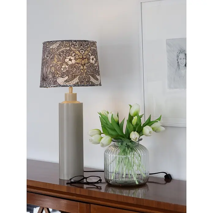 PR Home Tischleuchte Solo - 40 W, E27 - Eine graue Tischlampe mit gemustertem Vogel- und Blumenschirm auf einem Holzschrank neben weißen Tulpen und Brille.