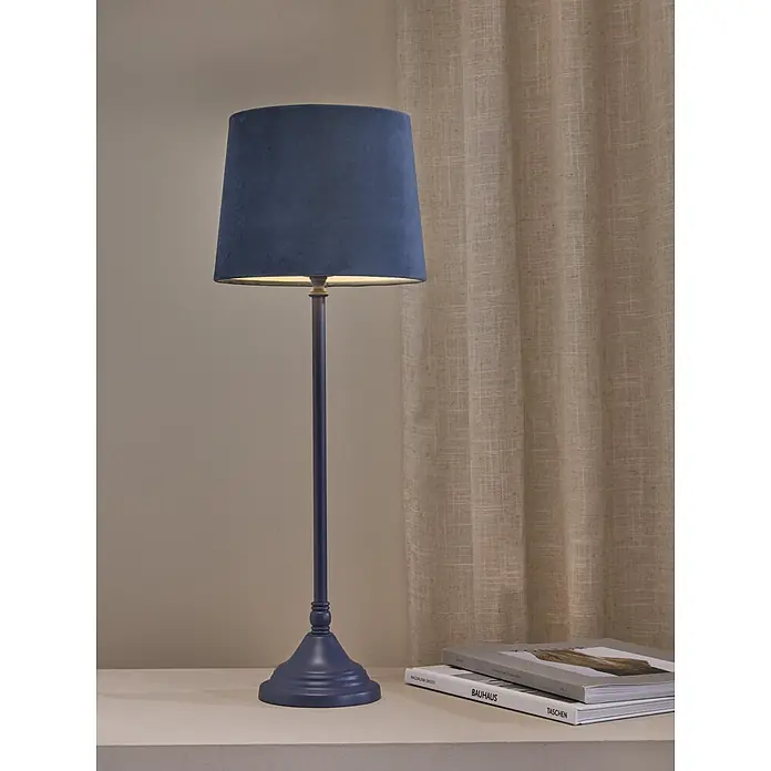 PR Home Tischleuchte Tove - 40 W, E27 - Dunkelblaue Stofftischleuchte mit Metallsockel auf einer Ablage, neben Büchern und einem Vorhang.