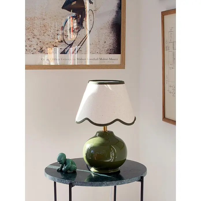 PR Home Tischleuchte Nell - 40 W, E27 - Grüne Keramiktischlampe mit cremefarbenem, gewelltem Lampenschirm und dunkler Borte, auf einem dunkelgrünen Marmortisch neben einer kleinen Figur.