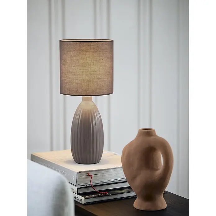 PR Home Tischleuchte Uno - 25 W, E14 - Gerippte Keramik-Tischlampe mit braunem Stoffschirm, beleuchtet Bücherstapel auf dunklem Tisch neben Terrakottavase.
