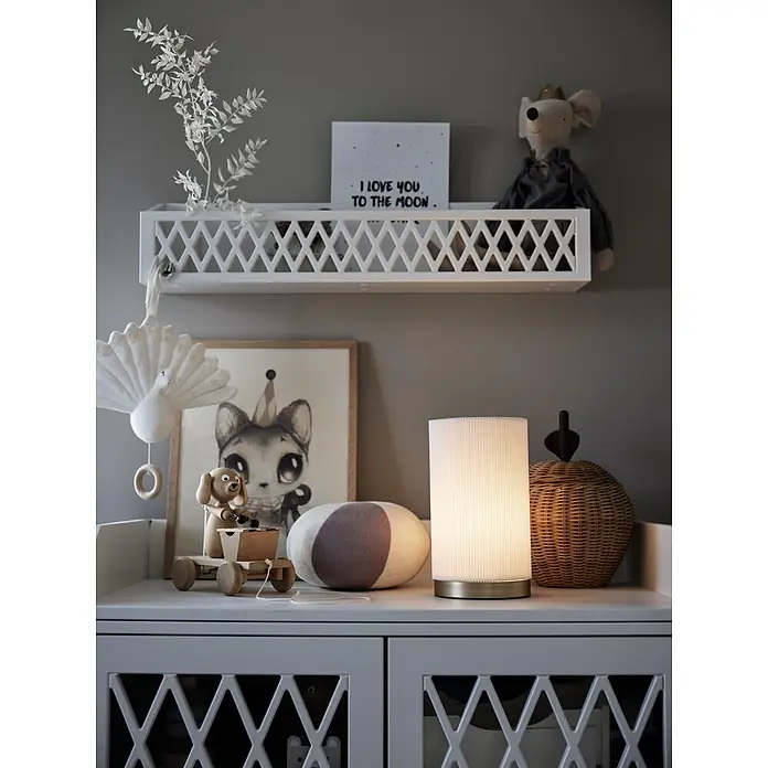 PR Home Tischleuchte Dora - 25 W, E27 - Ein weißes Regal mit geometrischem Gittermuster, das Kinderzimmer-Dekoartikel über einer Kommode mit Spielzeug und einer Lampe hält.