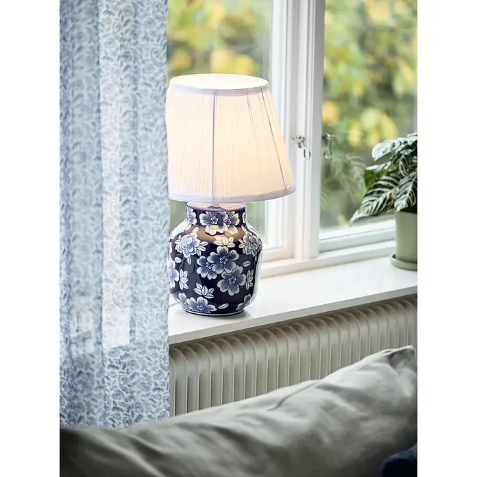 PR Home Tischleuchte Fanny - 15 W, E27 - Beleuchtete Tischlampe mit blau-weißem Keramikfuß mit Blumenmuster und plissiertem weißem Schirm auf einer Fensterbank.