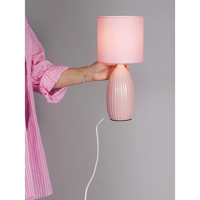 PR Home Tischleuchte Uno - 25 W, E14 - Rosa Tischlampe, von einer Hand in einem gestreiften Hemd gehalten, mit beleuchtetem Schirm und geripptem Fuß.