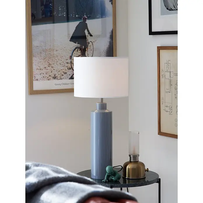 PR Home Tischleuchte Solo - 40 W, E27 - Eine blaue zylindrische Tischlampe mit weißem Schirm steht auf einem dunklen Beistelltisch. Eine Messinglaterne und eine grüne Figur sind ebenfalls auf dem Tisch.