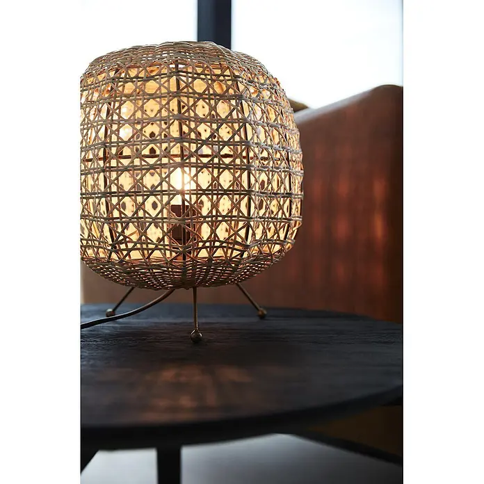 PR Home Tischleuchte Tischleuchte Wickham - 25 W, E27 - Eine beleuchtete, kugelförmige Tischlampe mit geflochtenem Rattanschirm auf einem dunklen Holztisch, die warmes Licht wirft.