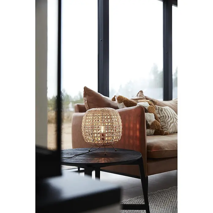 PR Home Tischleuchte Tischleuchte Wickham - 25 W, E27 - Kugelförmige Tischlampe aus geflochtenem Rattan leuchtet auf einem dunklen Beistelltisch neben einem braunen Sofa.