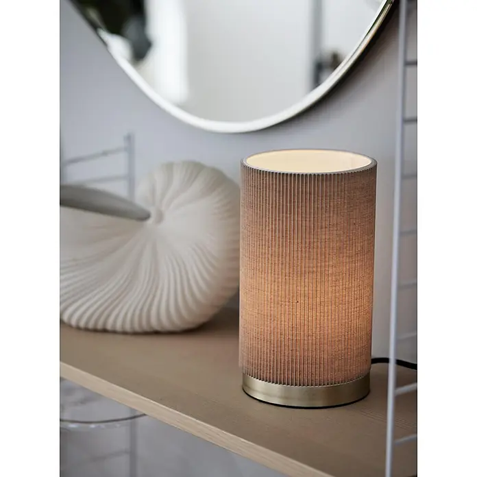 PR Home Tischleuchte Dora - 25 W, L x B x H: 14 x 14 x 25 cm, E27 - Zylindrische Tischlampe mit plissiertem beigem Stoffschirm und Messingsockel, leuchtend auf einem Holzregal.