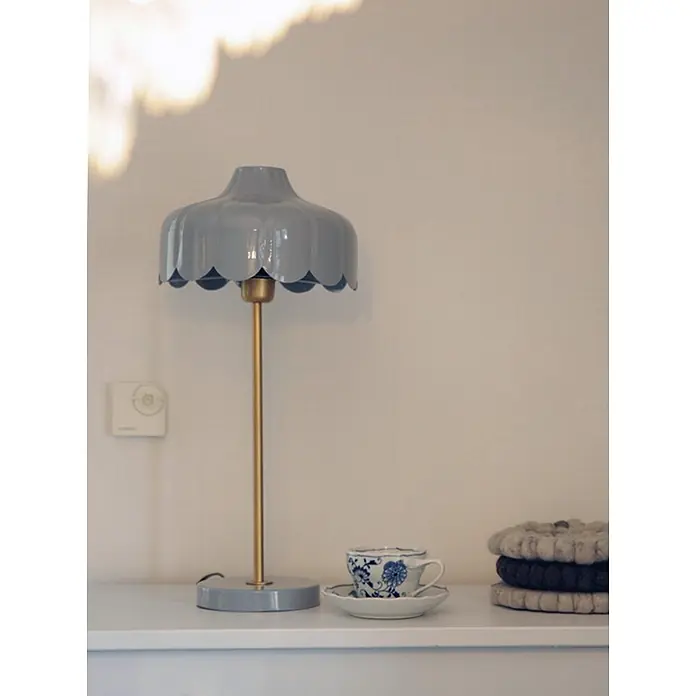 PR Home Tischleuchte Wells - 25 W, E27 - Eine hellgraue Tischlampe mit gewelltem Schirm und goldenem Ständer, daneben eine blau-weiße Teetasse und gestapelte Untersetzer.
